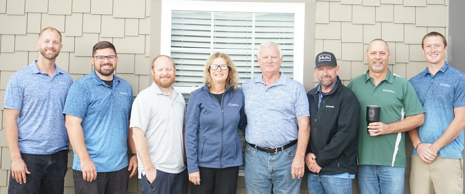 Eight members of the Triumph Construction team standing side by side in front of a shingle-sided building